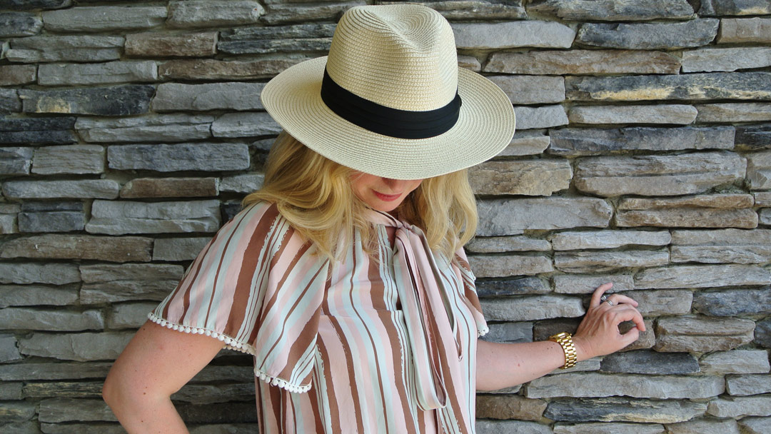 Laura wearing a paper straw Fedora hat looking down in front of a stone wall