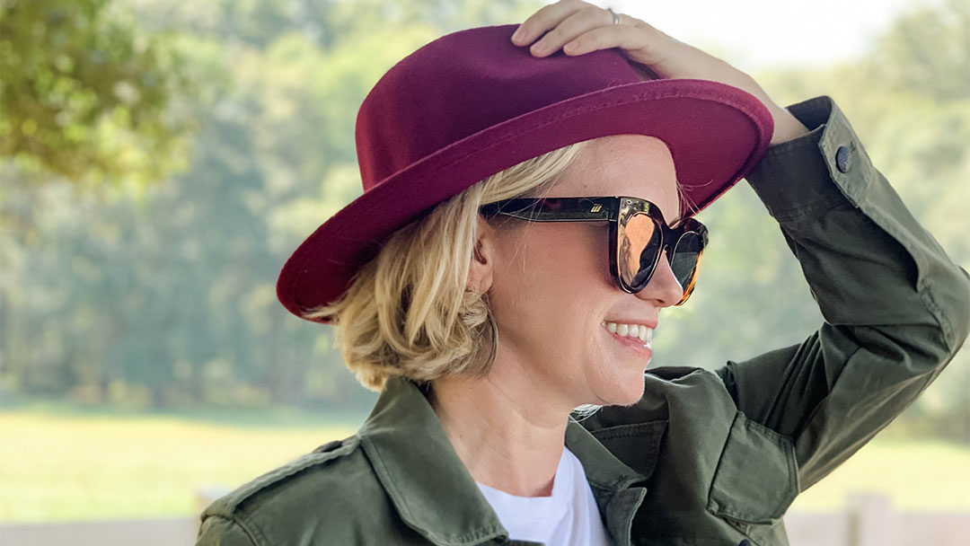 Side view of woman in sunglasses holding onto maroon hat