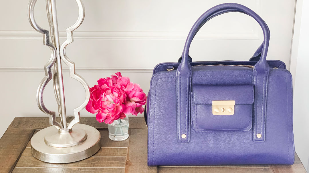Purple handbag on dresser with lamp and pink peony in vase